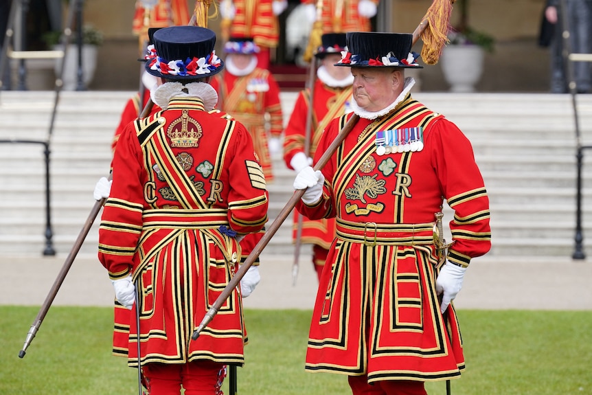 Men wearing traditional red uniforms and black hats are pictured carrying a spear.