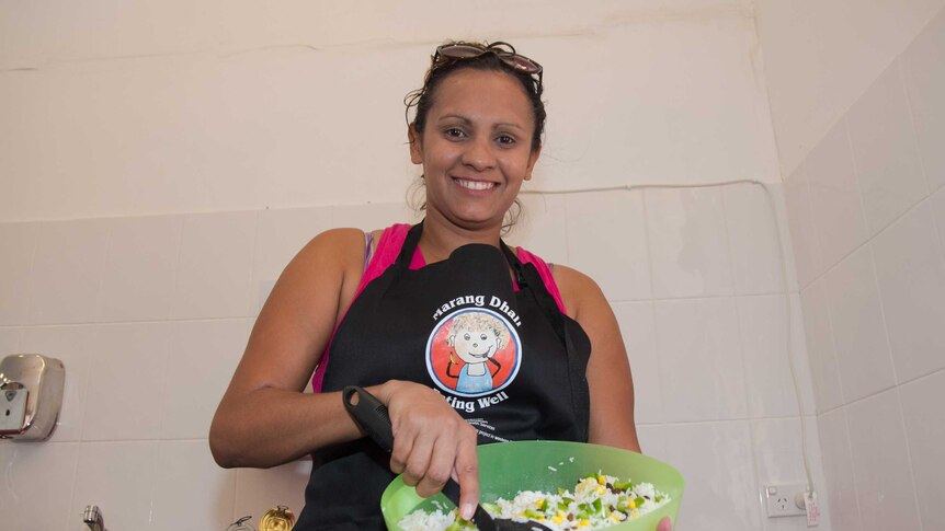 A woman in an apron saying Marang Dhali holding a bowl of rice salad