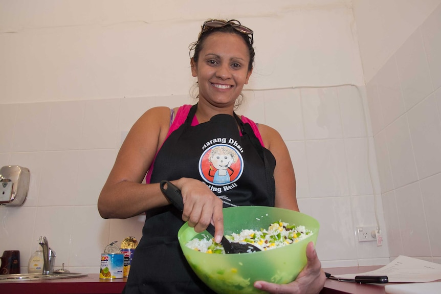 A woman in an apron saying Marang Dhali holding a bowl of rice salad