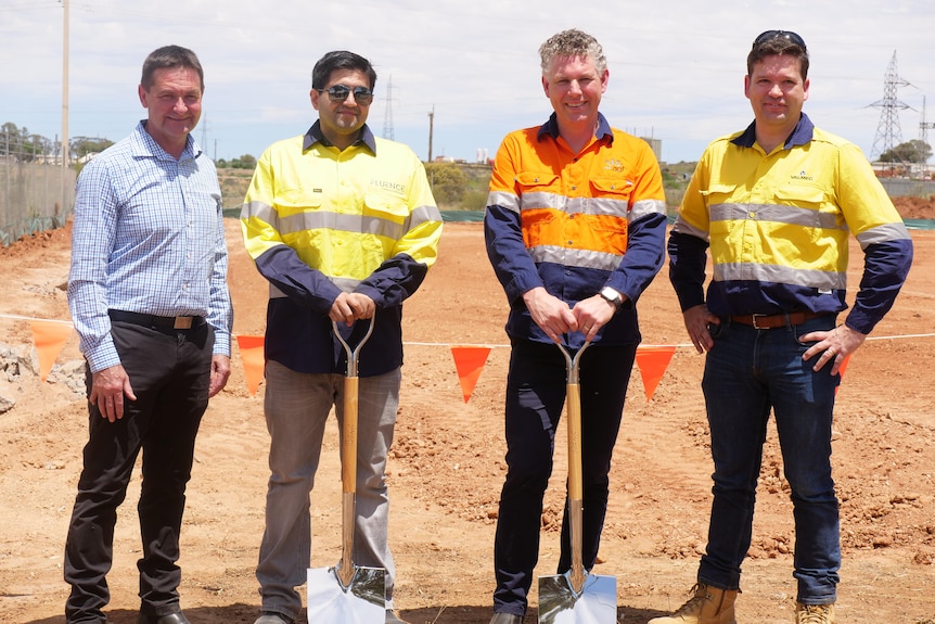 Four men standing in front of a construction site looking happy, two of whom are holding metal shovels