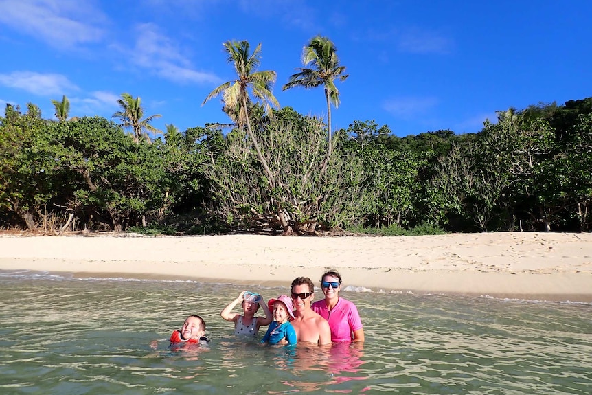 two adults and three children swim in shallow blue water with white beach and palm trees in the background