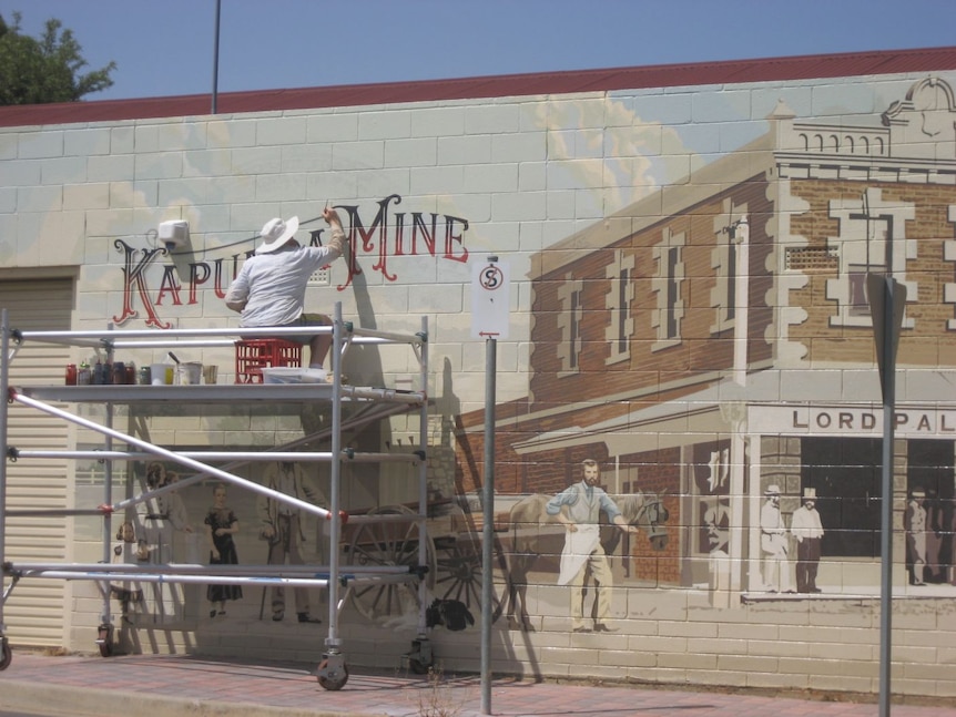 A man on scaffolding painting a mural with a Victorian brown and white building, man in Victorian clothing.