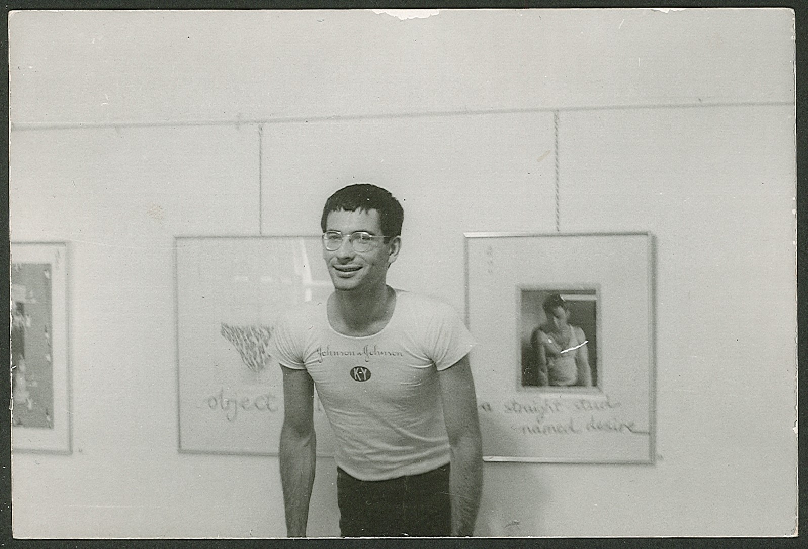 A black and white image of a white man with short dark hair and glasses, wearing a tight white t-shirt, standing in a gallery