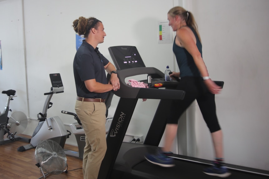 An exercise physiologist wearing pants and a polo shirt talks to a client, who is walking on a treadmill.