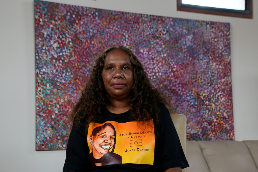 Bernadette sits in front of a dot painting, wearing a shirt that says stop Black Deaths in Custody