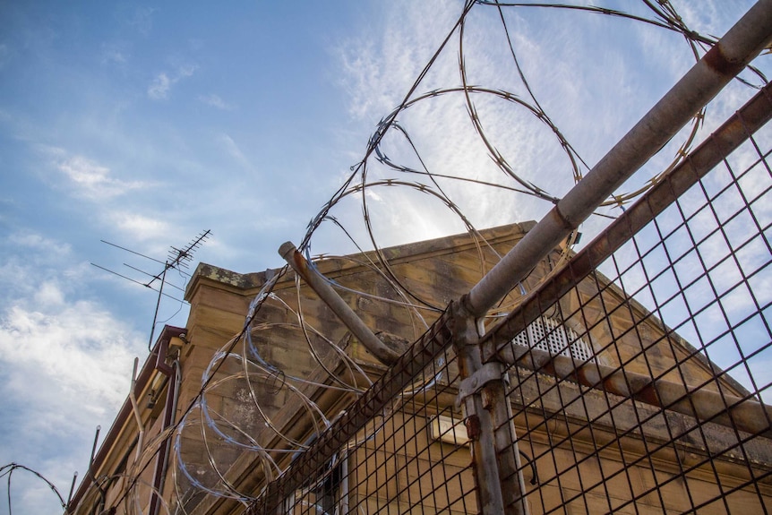 Razor wire sits atop a metal fence in front of a sandstone building.