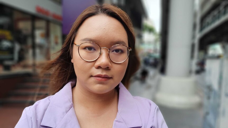 A young woman in a purple shirt and glasses stands on a street in Thailand