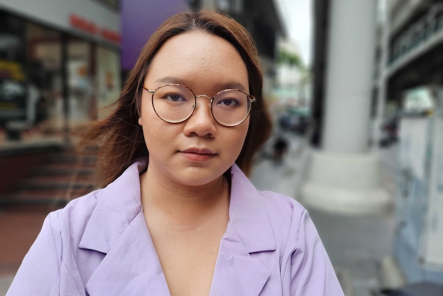 A young woman in a purple shirt and glasses stands on a street in Thailand