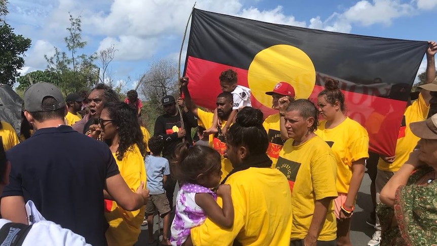 A number of Indigenous protesters wear yellow shirts and hold a large Aboriginal flag.