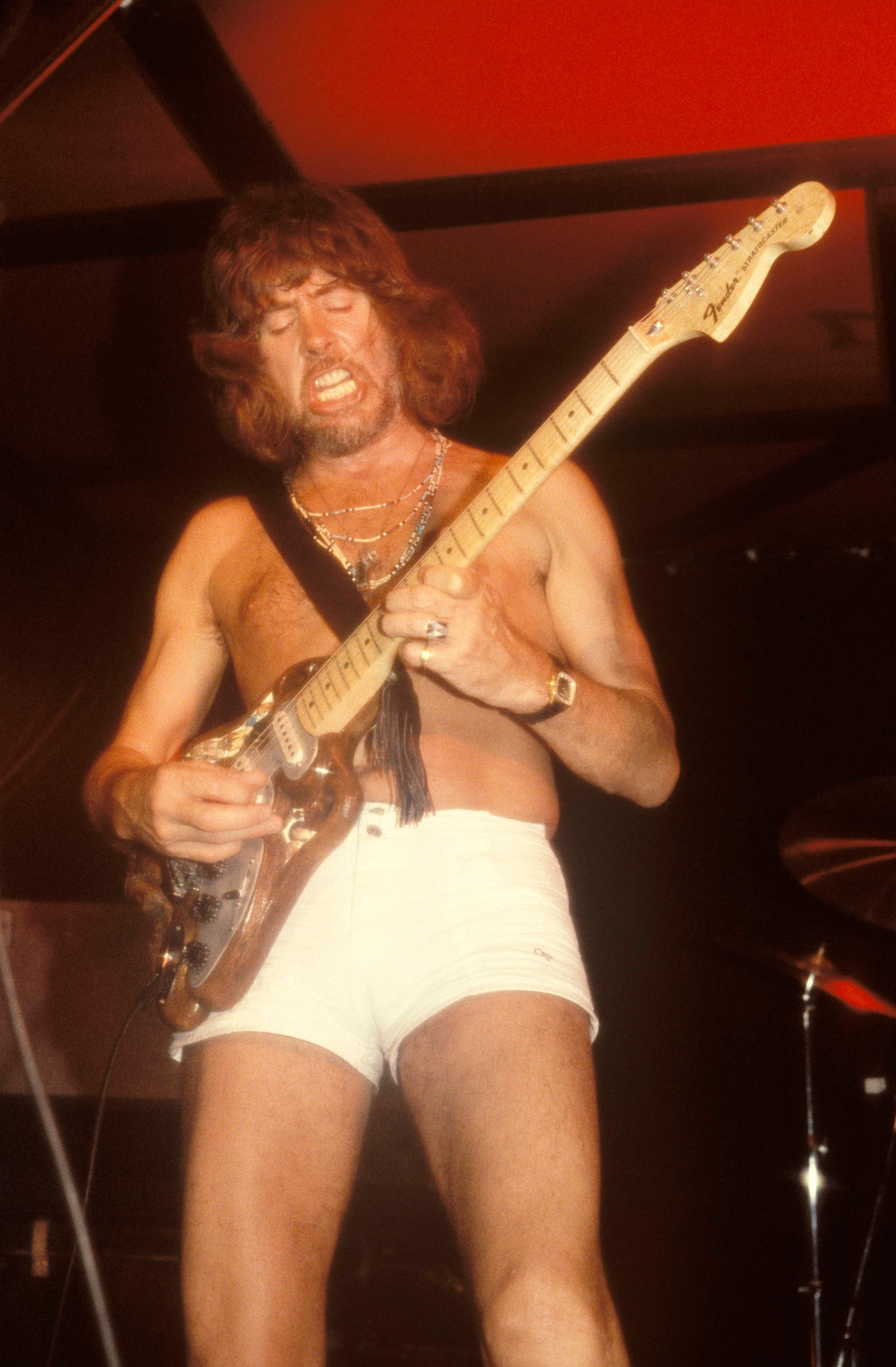 A shirtless John Mayall plays electric guitar on stage