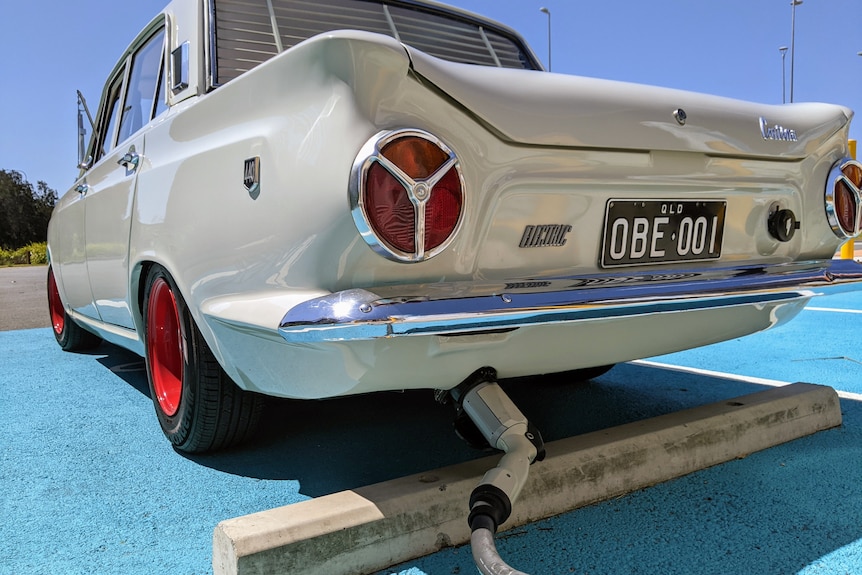 A low-angle shot of an old-fashioned white car with a cable plugged into a socket at the back