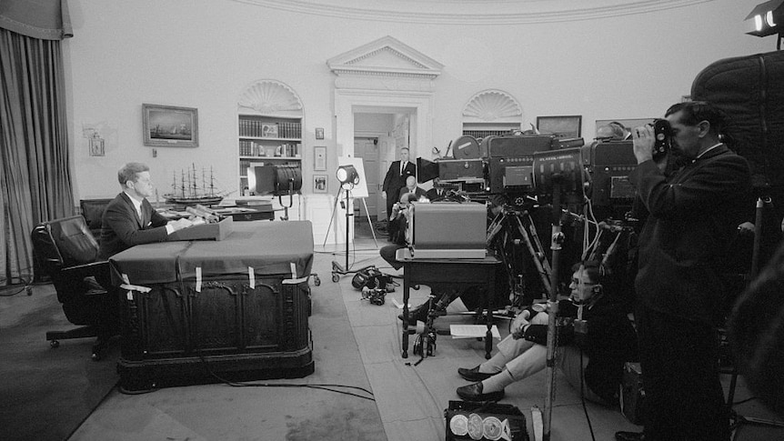 Black and white photograph of John F Kennedy behind a large desk in the Oval Office addressing several journalists and cameras