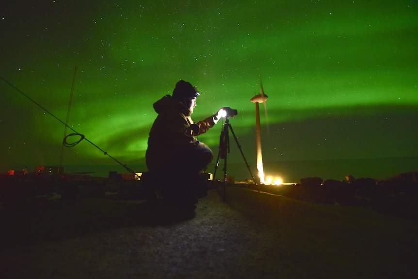 A man crouches in front of a camera at night with the green glow or Aurora Australis around him