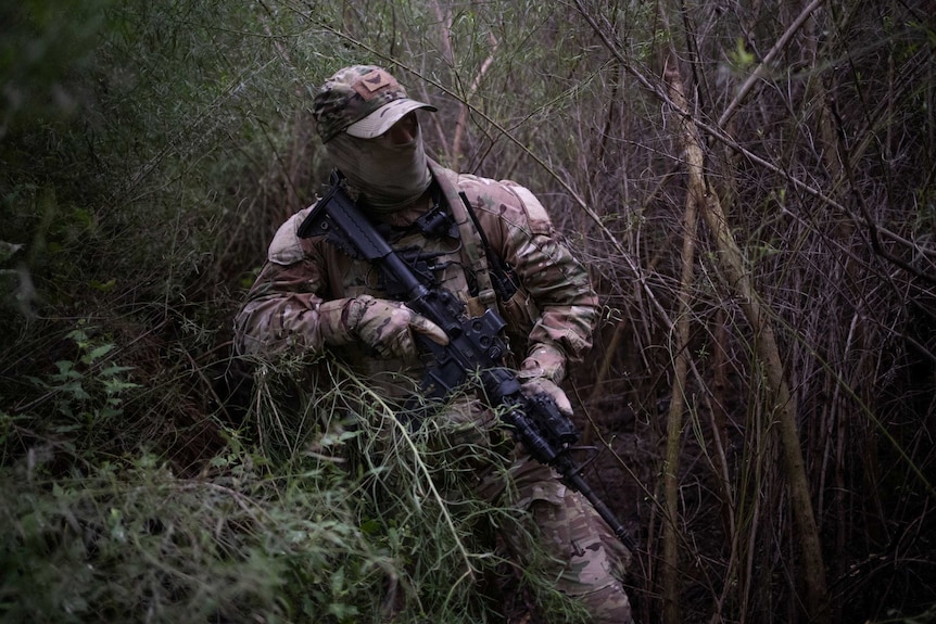 A man in heavy camouflage and a gun in a wooded area