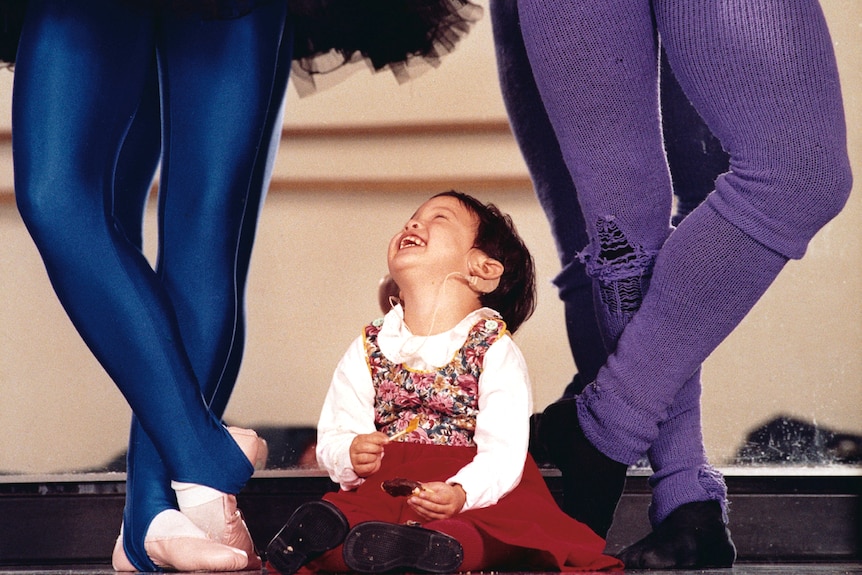 A little girl sits on the floor looking up at her parents in dance tights, only their legs can be seen