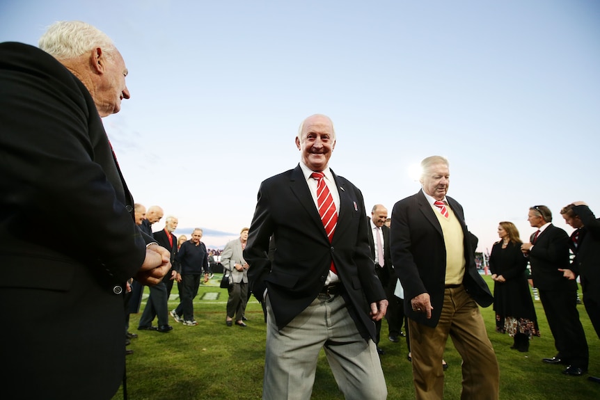 In a suit and red and white tie, Johnny Raper walks off a footy field