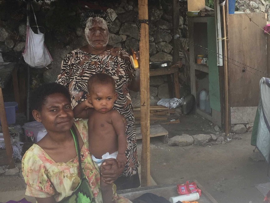 A woman sits on a bench inside a tent, holding her infant child on her hip. Another woman stands behind them.