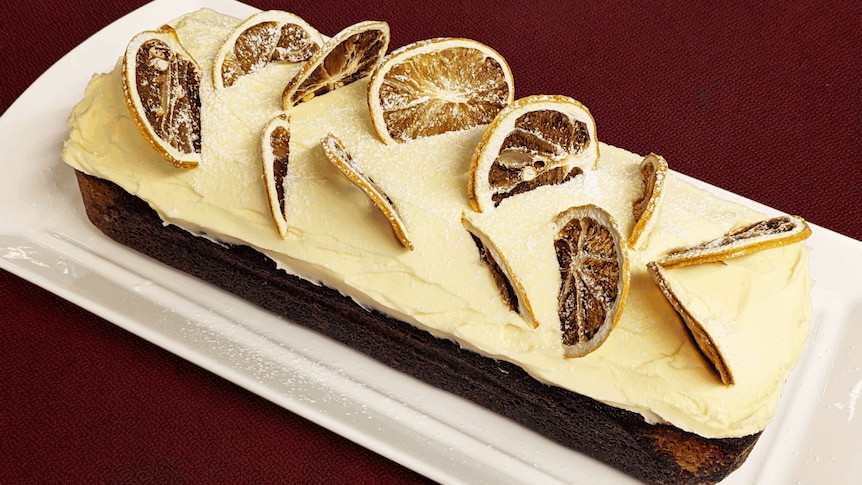 Loaf-shaped cake on a plate, topped with pale icing and dried citrus slices.