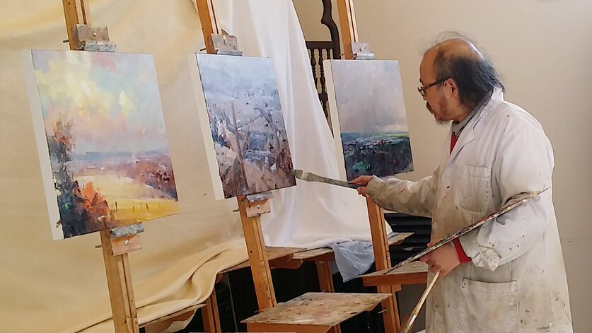 An artist with a brush and palette of paints stands poised over three square canvases.