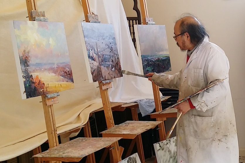 An artist with a brush and palette of paints stands poised over three square canvases.