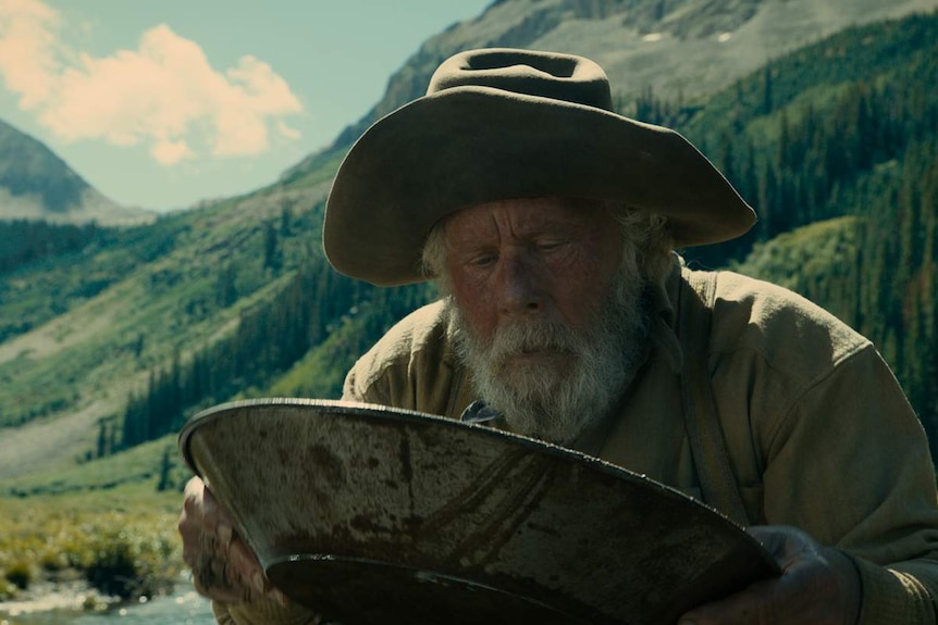 Old man with grizzled beard and battered hat holding prospecting pan, river and green-treed mountains in background.