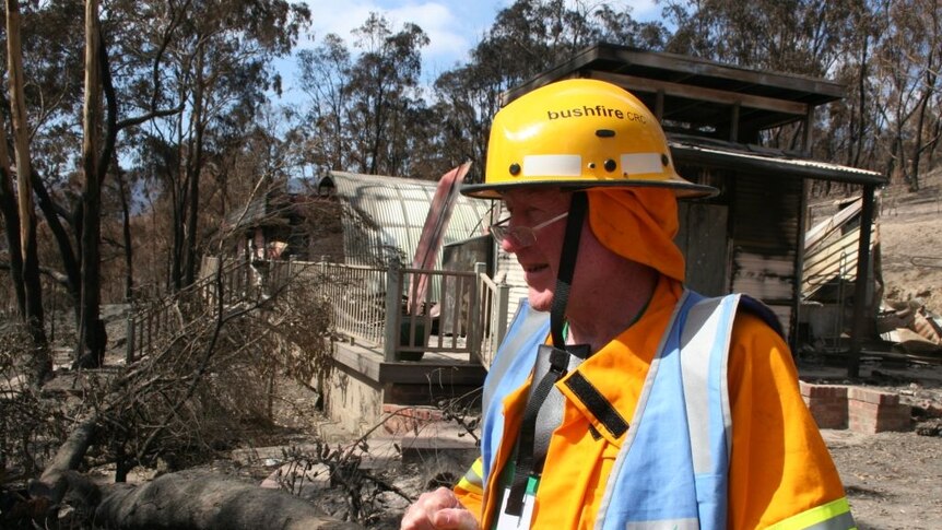 Dr Jim McLennan in firefighting gear stands on a property that has recently been burned. He is wearing glasses.