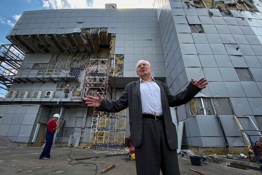 An image of a man wearing a suit standing in front of a damaged grey building with scaffolding in front of it. 