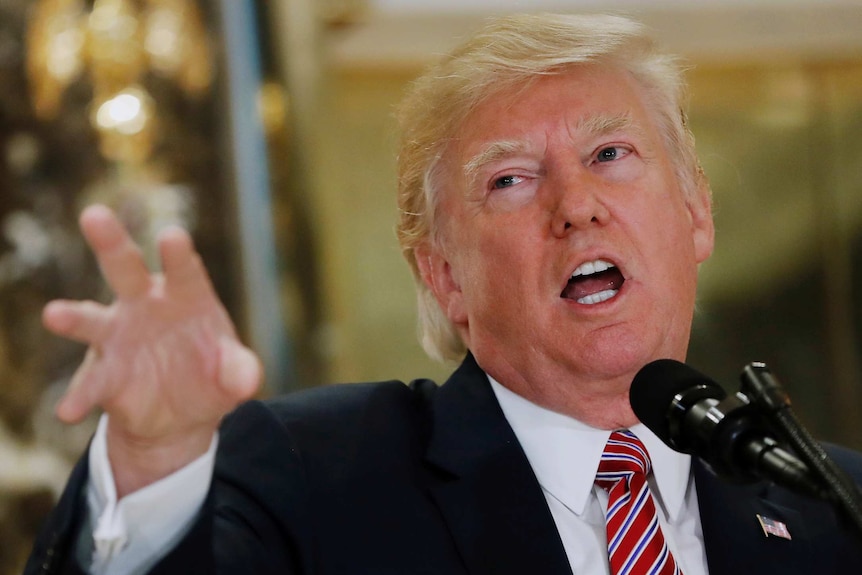 US President Donald Trump speaks at microphone while gesturing with his right hand. Face is red.