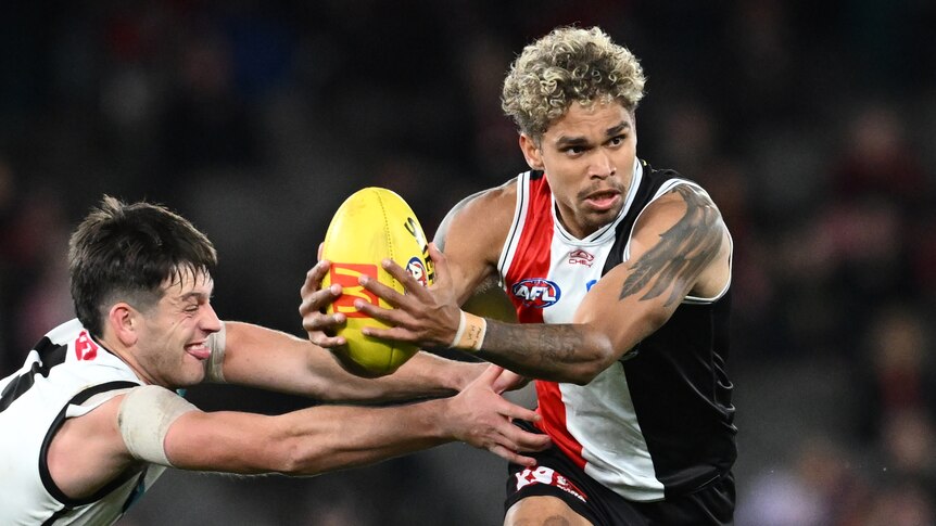 A St Kilda AFL player holds the ball and looks downfield as a Port Adelaide player tries to hang on to him from the side.
