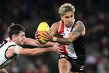 A St Kilda AFL player holds the ball and looks downfield as a Port Adelaide player tries to hang on to him from the side.
