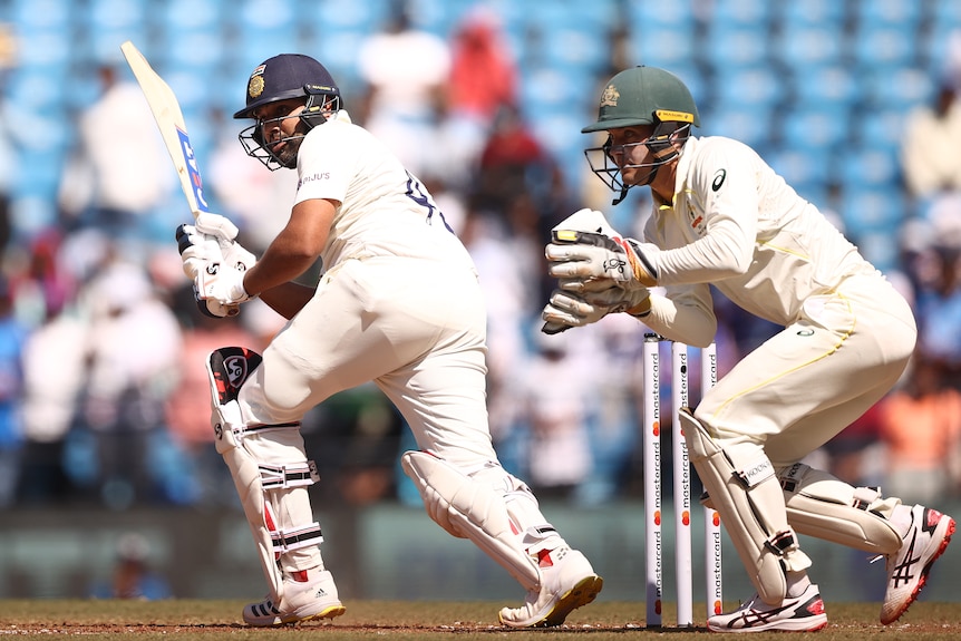 An Indian male batter plays a shot to the leg side as the Australian wicketkeeper looks on.