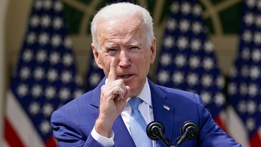 A white-haired President Joe Biden wags his finger during a speech. He has two crossed American flags behind him