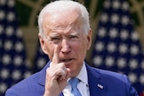 A white-haired President Joe Biden wags his finger during a speech. He has two crossed American flags behind him