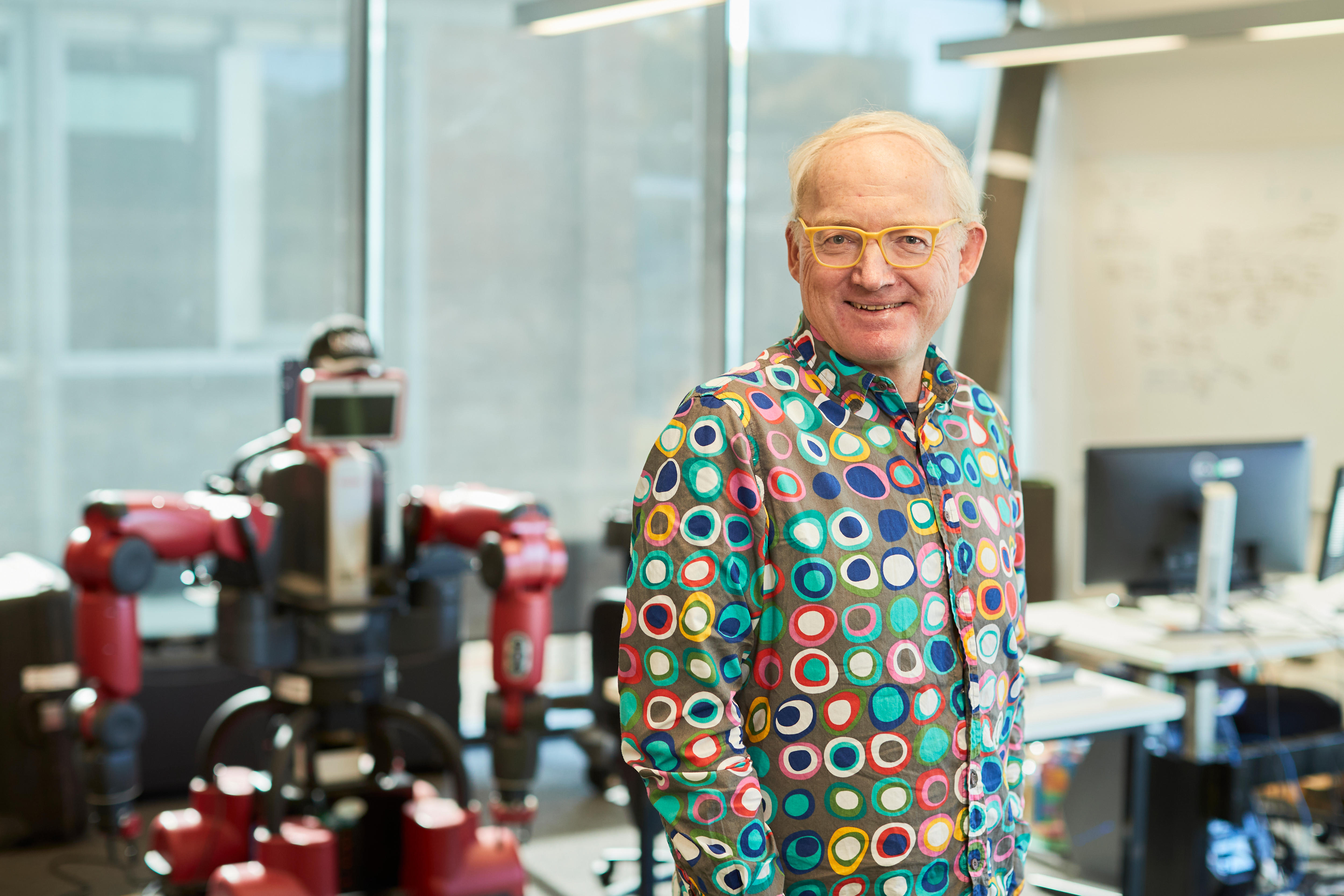 Um homem com uma camisa colorida e óculos fica na frente de um robô.