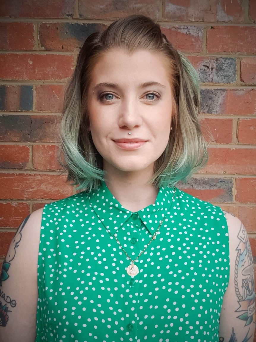 Erika is wearing a pale green top and standing in front of a brick wall. She has green-tinted hair and have a lip piercing.