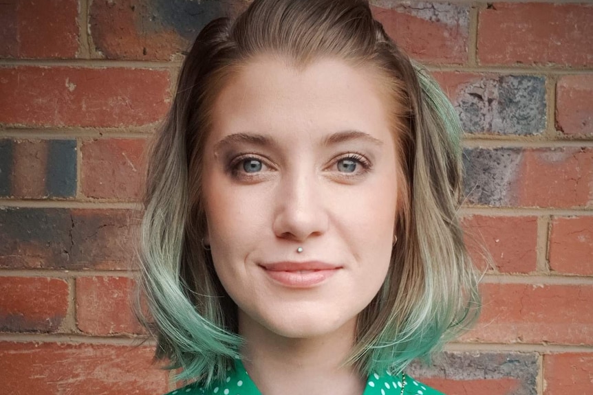 Erika is wearing a pale green top and standing in front of a brick wall. She has green-tinted hair and have a lip piercing.