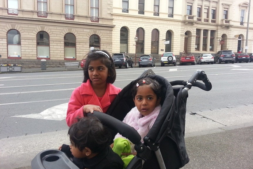 Two young children in a pram and one stands behind on a city street with a building in the background.