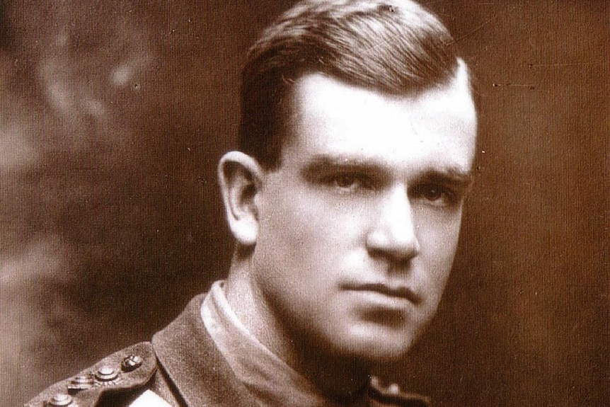 An black and white photo of a man in his mid-20s, standing side on, arms folded, he is wearing a military jacket.