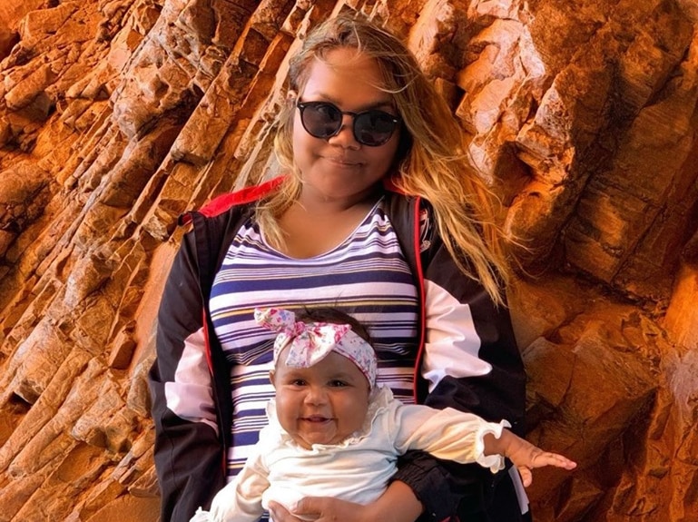 Photo of an Aboriginal woman in front of a red rock face holding an Aboriginal baby