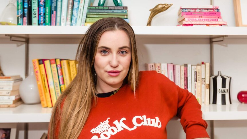 Lina Abascal is an American writer and she is standing in a red jumper in front of a bookcase