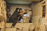 Two people, a man and a woman, sit on a ledge in a cramped bathroom space, both looking at their phones
