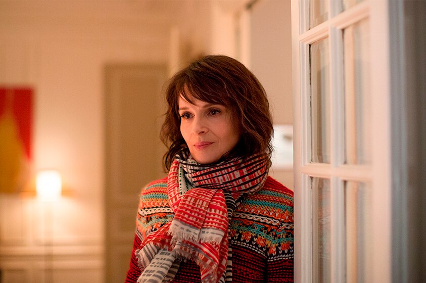 A brunette woman in bright patterned scarf and top, stands in doorway of warmly lit room with wistful expression.