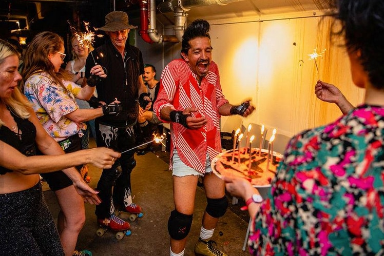 A man on roller skates stands with an excited look on his face as a birthday cake is carried towards him.