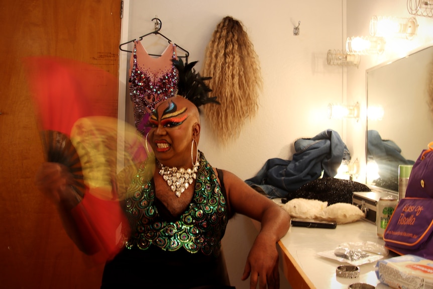 A man in drag is fanning himself, sitting at a dressing table. 
