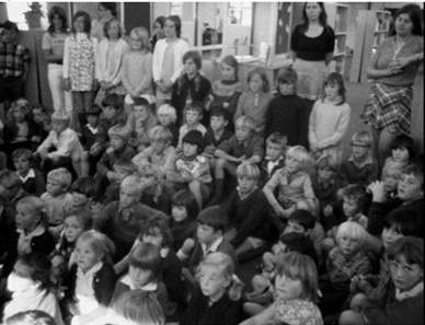 Black and white photo of children sitting on floor and two teachers standing at the back watching, some children stand also.