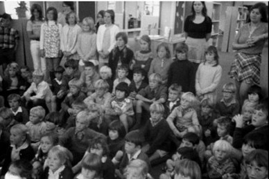 Black and white photo of children sitting on floor and two teachers standing at the back watching, some children stand also.