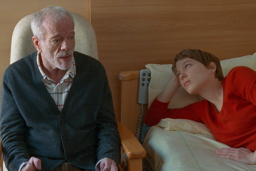 An older man sits on a chair beside a bed. A young woman lays on the bed resting her head on her arm, looking at the man.