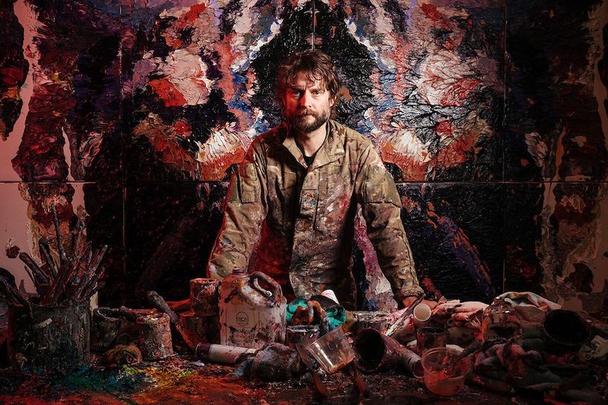 Australian artist Ben Quilty stands over a desk covered in panting supplies.