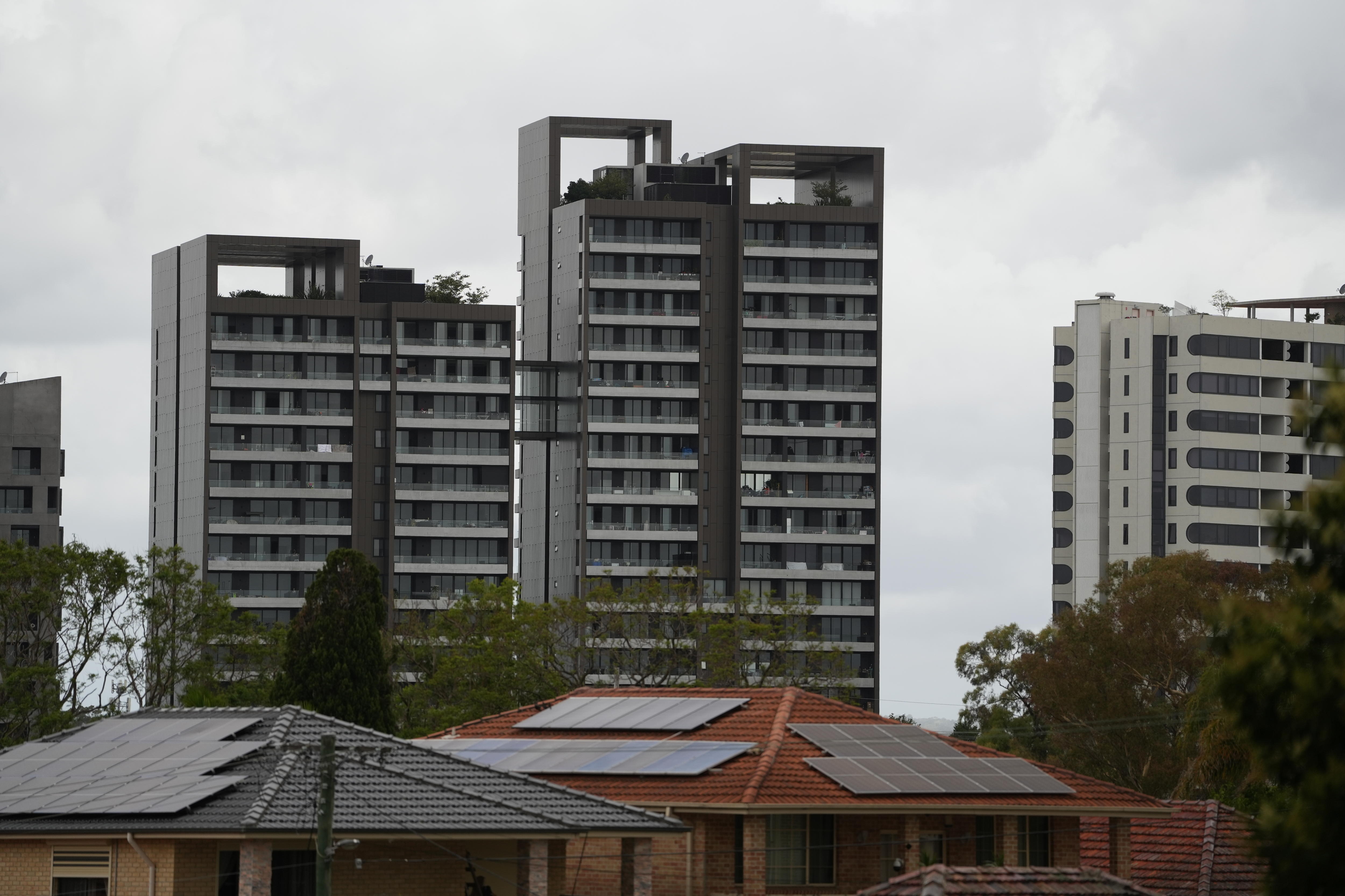 Tres nuevos edificios de gran altura, en primer plano casas con paneles solares bajo un cielo gris