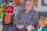 A man in glasses sitting on a couch, smiling next to four colourful metre-tall crochet characters.
